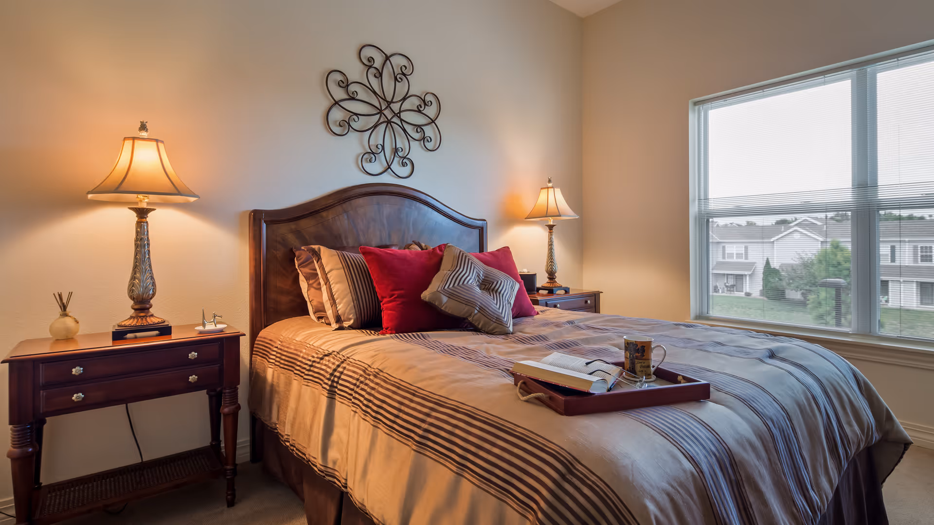 Well-lit bedroom with a neatly made bed topped with decorative pillows, two bedside tables with lamps, and a large window overlooking neighboring buildings.
