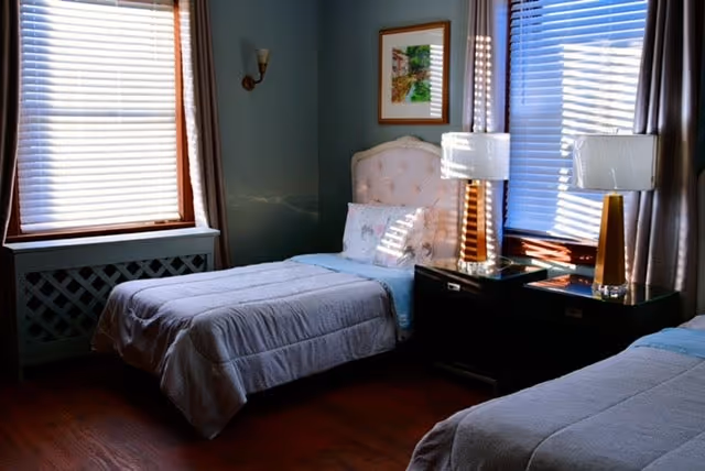 A cozy bedroom with two single beds, each with light-colored bedding. Between the beds is a dark nightstand with two modern table lamps. The room has wooden floors, two large windows with blinds and curtains, and a framed picture on the wall.