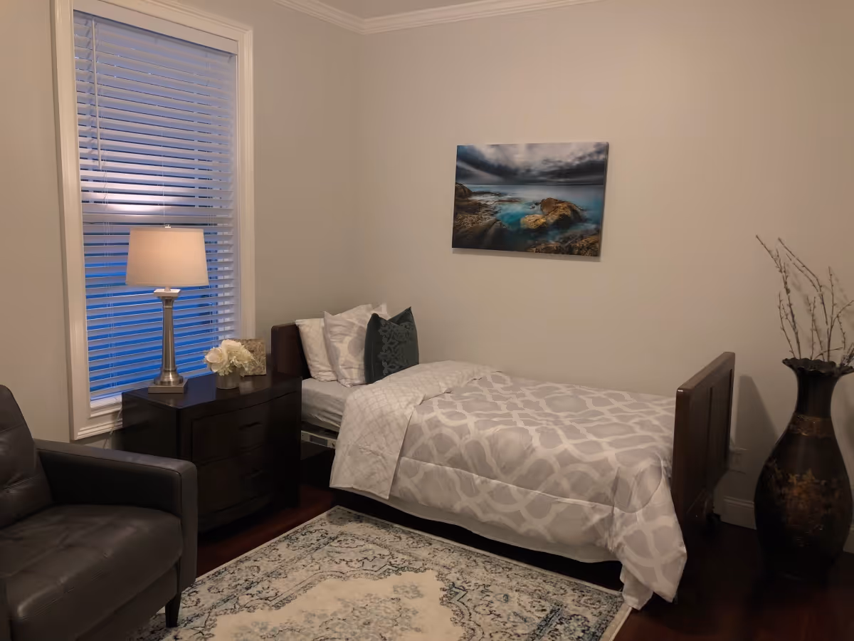 A cozy bedroom with a single bed covered in a white and gray patterned comforter and pillows. Next to the bed is a dark wooden nightstand with a lamp, a small flower arrangement, and a decorative item. A large window with white blinds is behind the nightstand. There is a dark leather armchair in the corner, a decorative vase with branches on the floor, and a framed picture of a rocky seashore hanging on the wall above the bed. The room has light-colored walls and a patterned area rug on the wooden floor.
