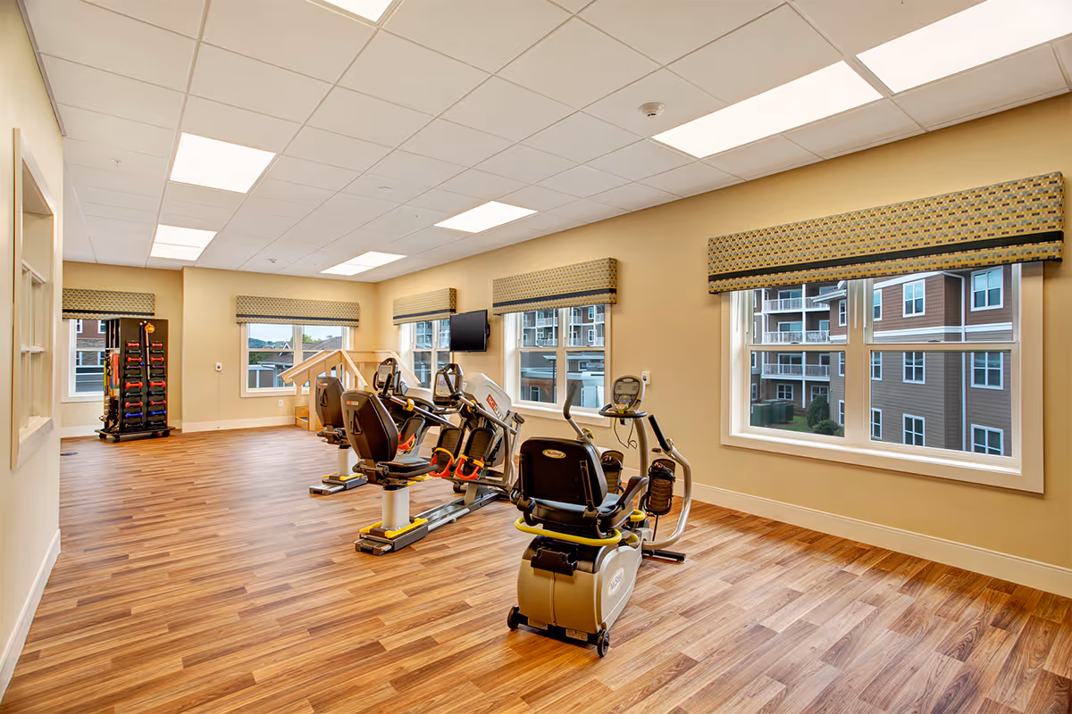 Well-lit fitness room with several recumbent exercise bikes, a rack of free weights, and large windows overlooking neighboring apartments.