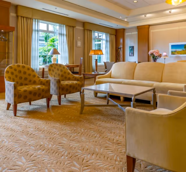 A cozy living room area with a beige sofa, two patterned armchairs, a glass-top coffee table, and a beige carpet with a leaf pattern. The room has large windows with curtains, table lamps, and decorative flowers on a side table, creating a warm and inviting atmosphere.