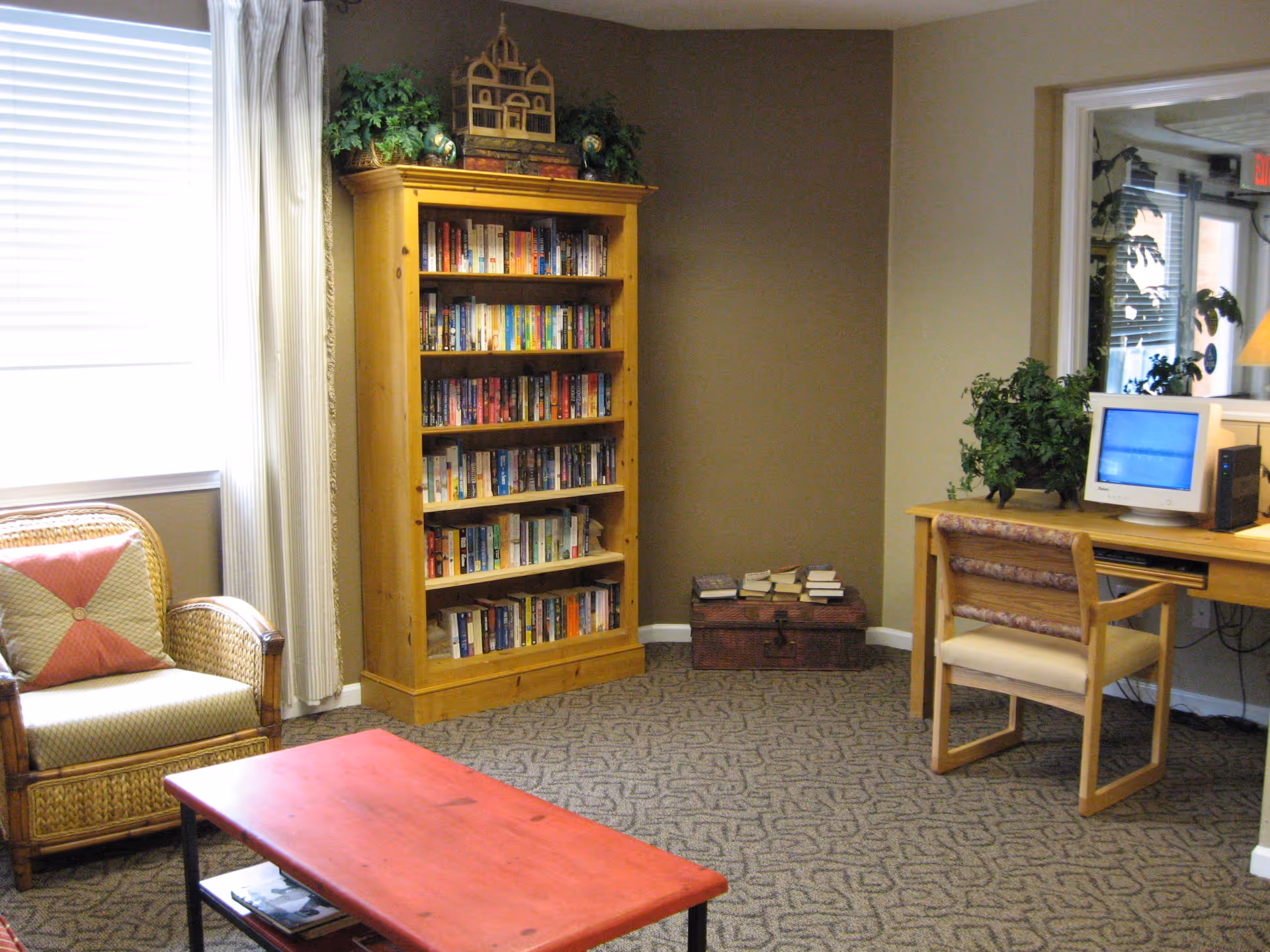 A cozy room with a wicker armchair with a patterned cushion, a red wooden coffee table, a tall wooden bookshelf filled with books, and a wooden desk with a computer and a chair. There are plants on top of the bookshelf and desk, and a window with white curtains letting in natural light.