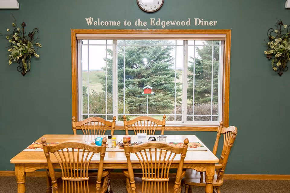 A dining area with a wooden table and six matching wooden chairs. The table is set with placemats and condiments. Behind the table is a large window showing green trees outside. Above the window, a sign reads 'Welcome to the Edgewood Diner'. The walls are painted green and decorated with floral wall hangings on either side of the window.