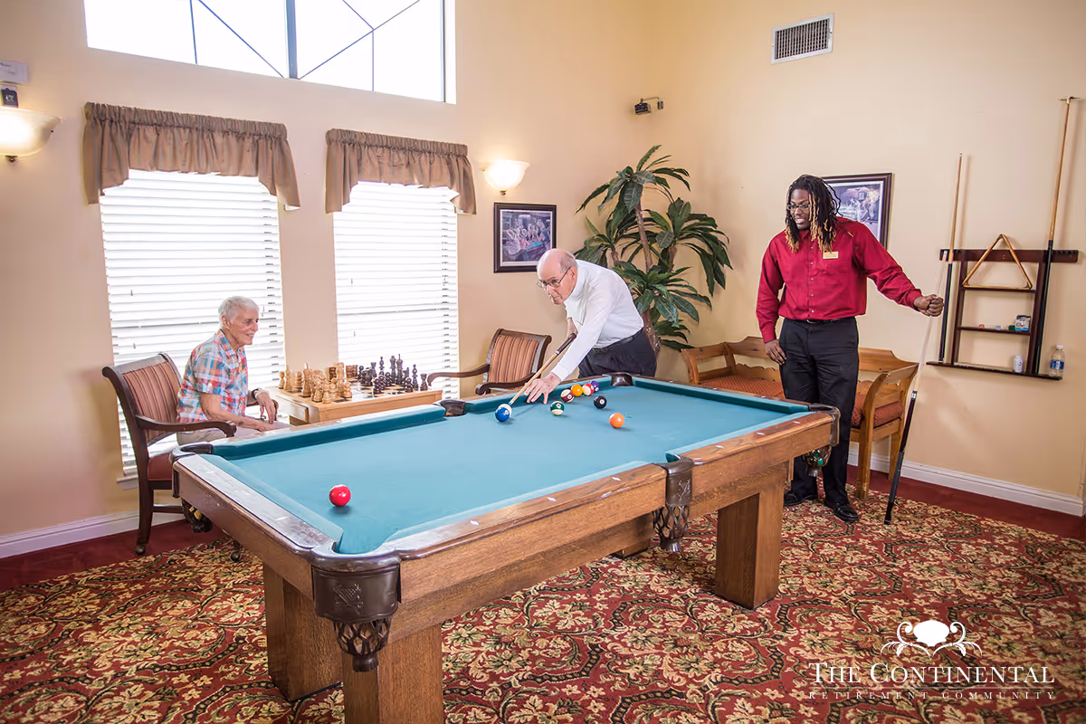 Residents play pool in a carpeted game room with a chess set by the windows and a staff member standing nearby.