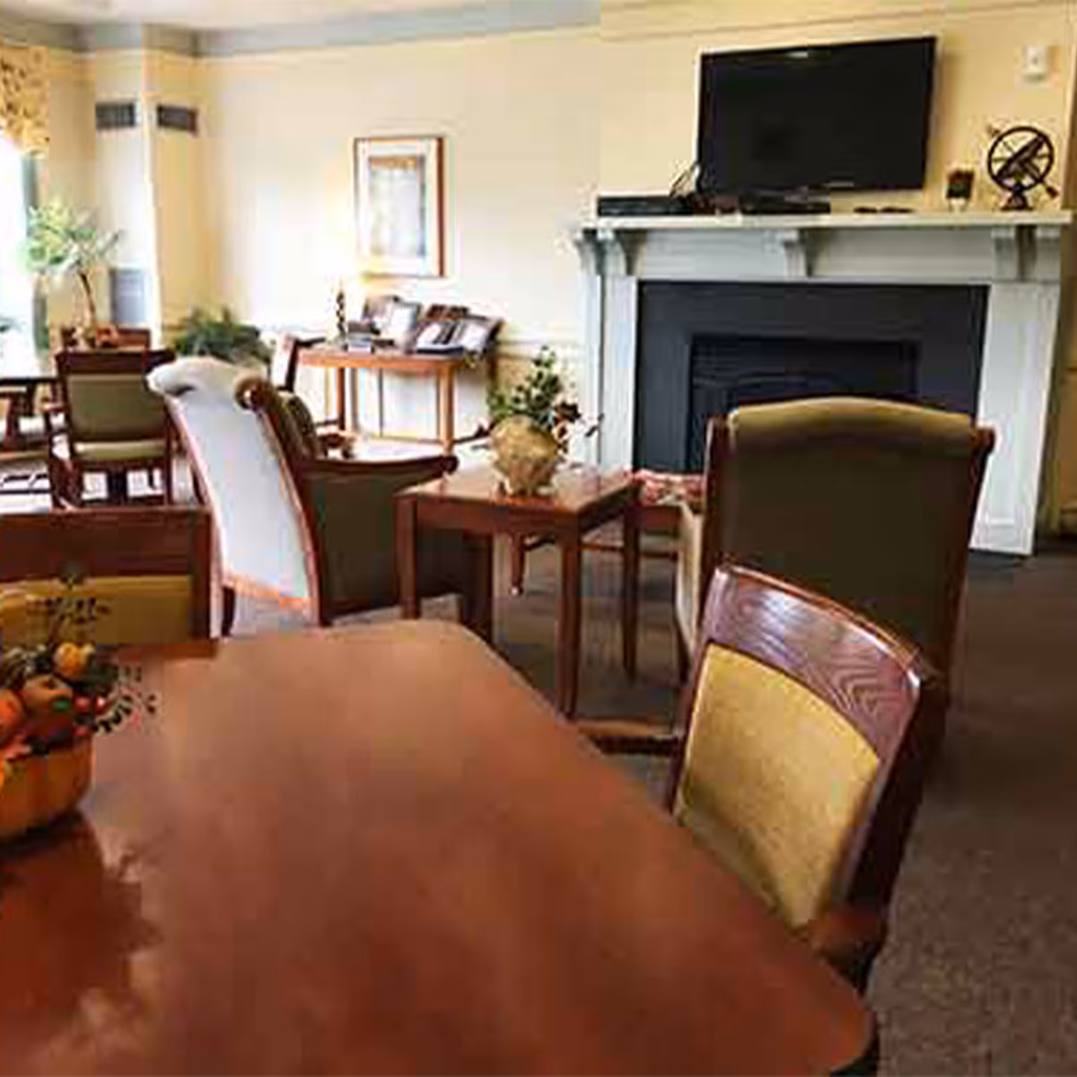 A cozy living room area with multiple wooden tables and upholstered chairs arranged around a fireplace with a flat-screen TV mounted above it. There are decorative plants and framed artwork on the walls, creating a warm and inviting atmosphere.
