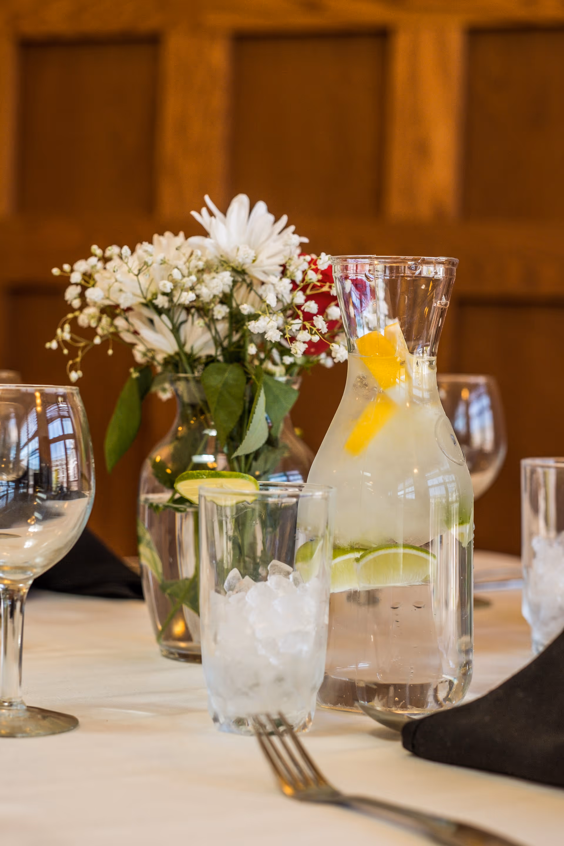 A table set with a carafe of lemon-and-lime water, an ice-filled glass, wine glasses, and a vase of white flowers.