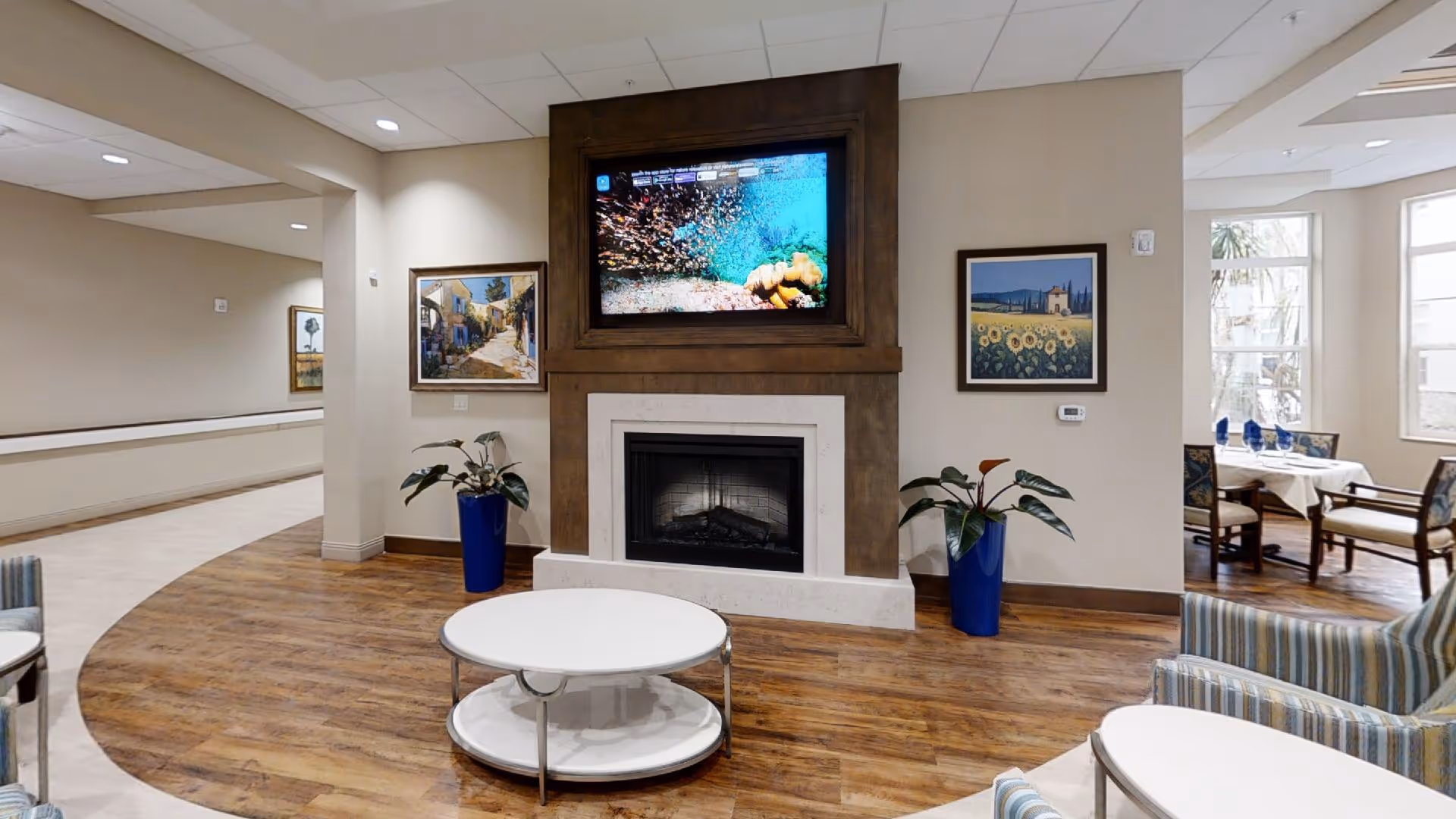 A cozy living room area with a fireplace and a large flat-screen TV mounted above it displaying an underwater scene. The room has wooden flooring, two blue vases with plants on either side of the fireplace, framed paintings on the walls, and a round white coffee table in the center. To the right, there is a dining area with tables and chairs near large windows letting in natural light.