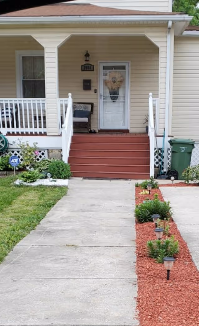 Concrete walkway leading to a beige house's covered front porch with red steps, white railings, a wreath on the door, and planted mulch beds with pathway lights.