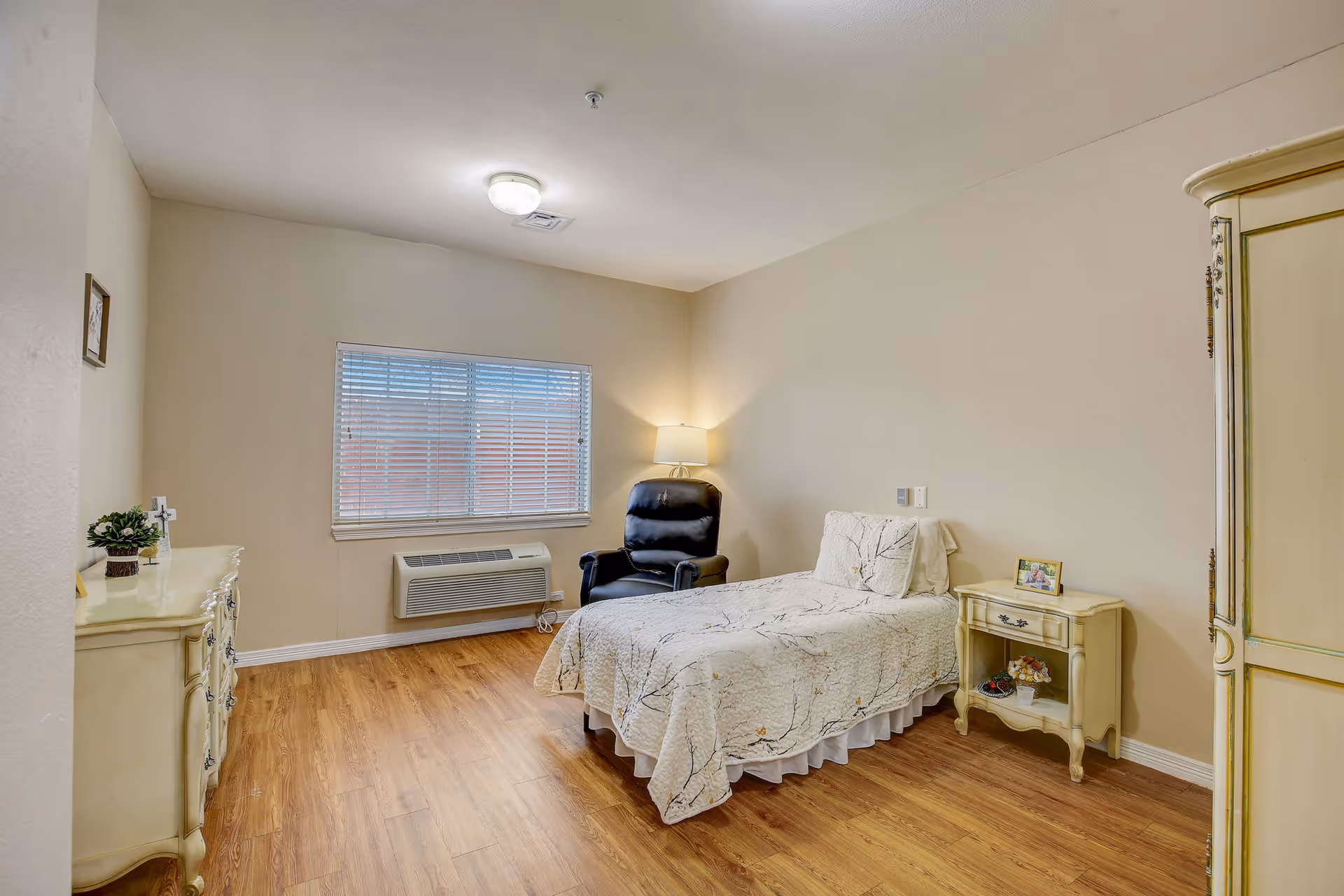 A simple bedroom with a single bed covered in a white quilt with a branch pattern, a black recliner chair next to a floor lamp, a window with blinds, a cream-colored dresser with a small plant and cross on top, and a matching nightstand with a framed photo and decorative items. The room has wooden flooring and beige walls.
