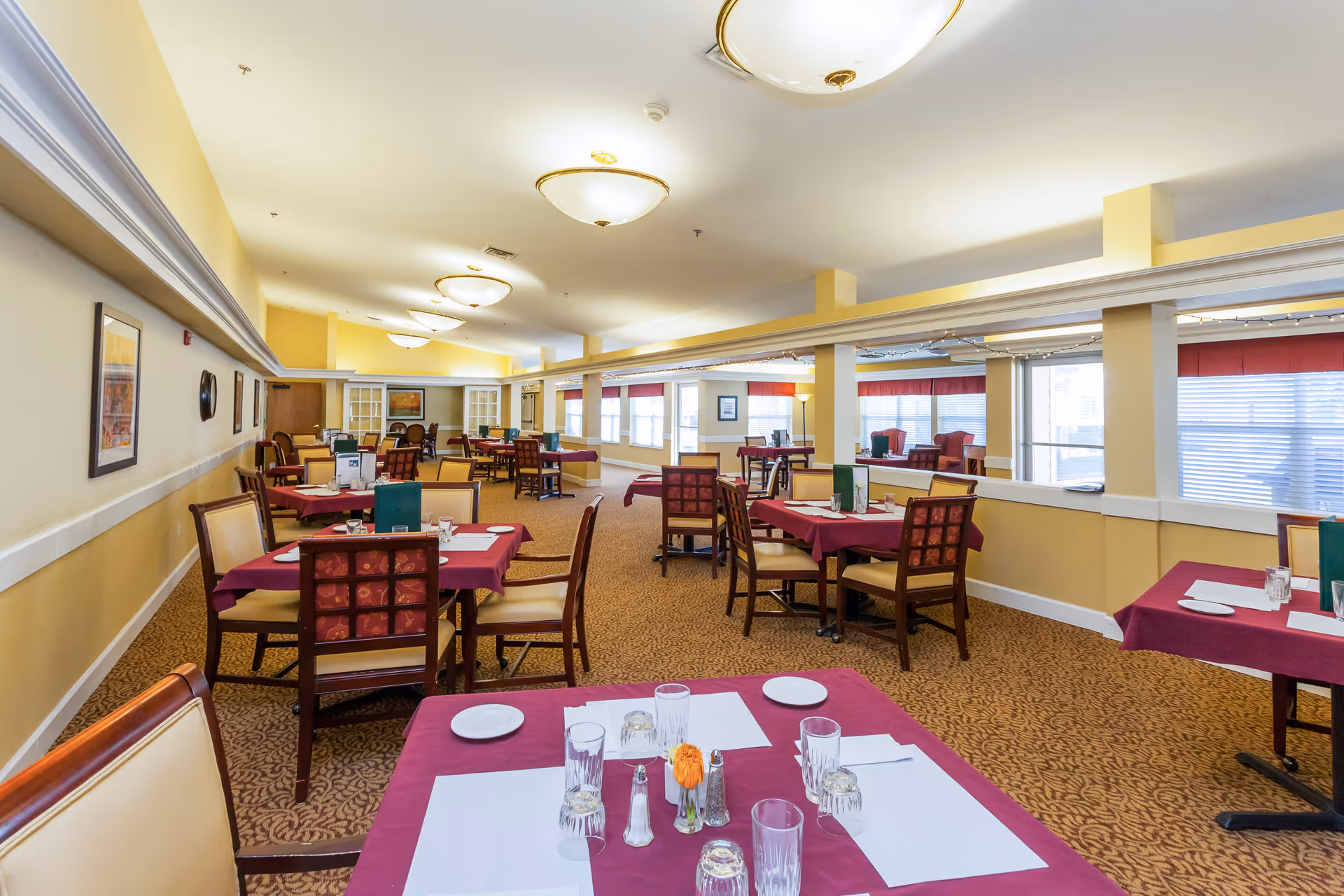 A spacious dining room in Lassen House Senior Living with multiple tables covered in burgundy tablecloths, each set with placemats, glasses, plates, and silverware. The room has beige walls, patterned carpet, and large windows with blinds allowing natural light. Ceiling lights illuminate the room, and framed artwork decorates the walls.