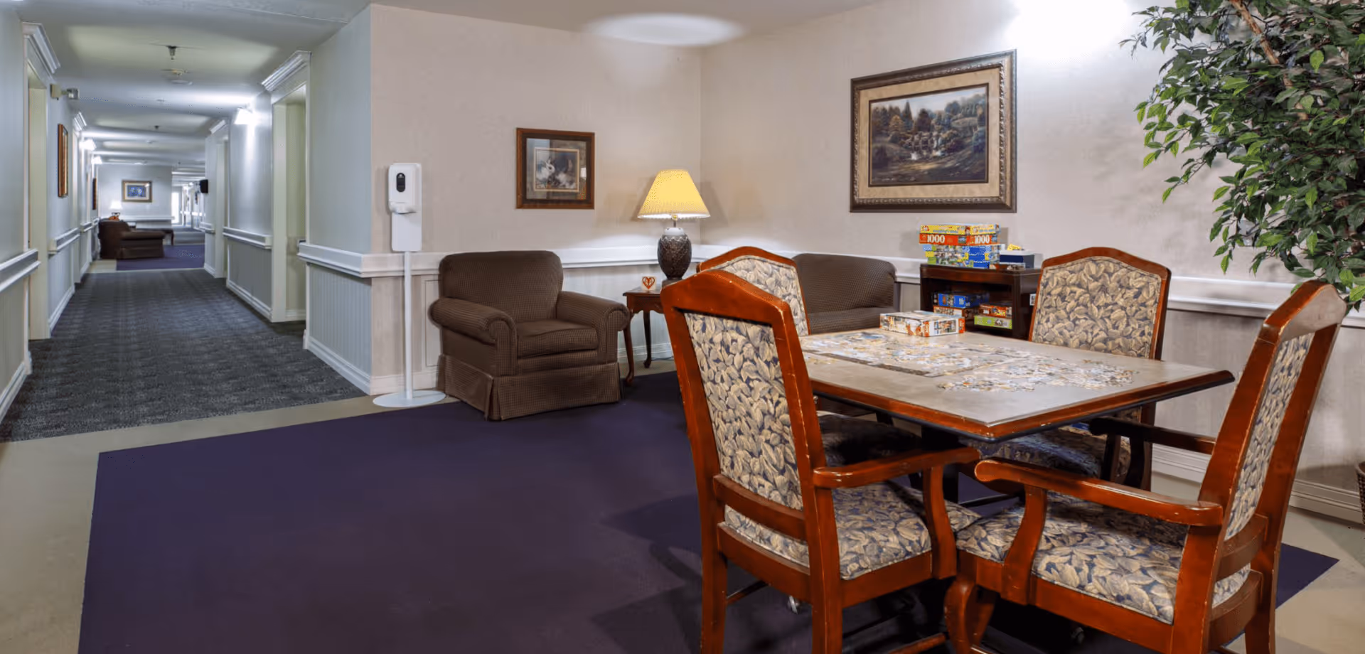A cozy seating area in a senior living facility hallway with a table and four cushioned chairs arranged around it. The table has a partially completed jigsaw puzzle on it. Behind the table, there is a brown armchair, a side table with a lamp, and a shelf with board games. The walls are decorated with framed paintings, and there is a large artificial plant on the right side. The hallway extends into the distance with more seating visible.