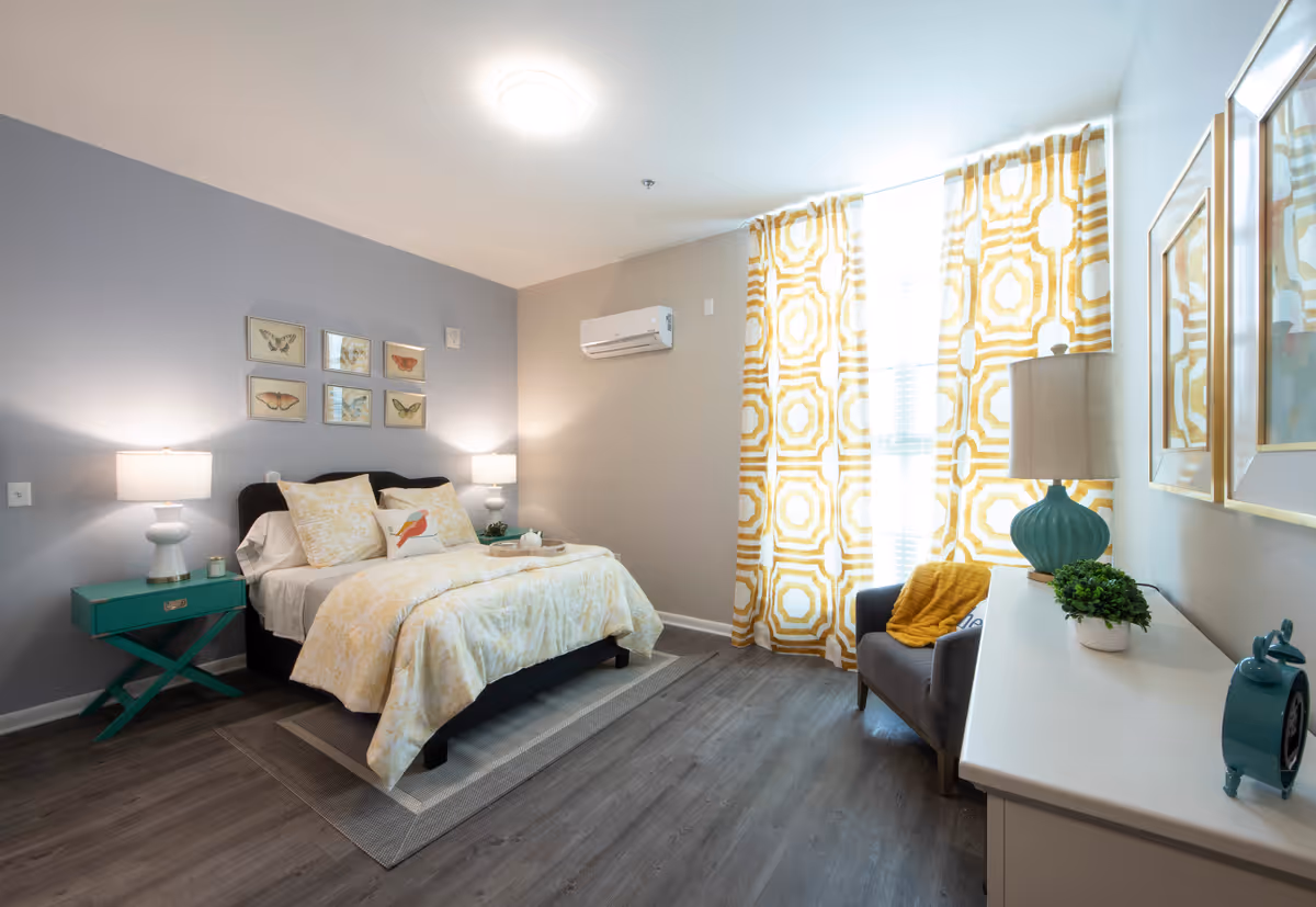 A bright and cozy bedroom with a bed dressed in yellow and white floral bedding, two bedside tables with lamps, butterfly artwork on the wall, a gray armchair with a yellow throw blanket, patterned yellow and white curtains, and a white dresser with decorative items.