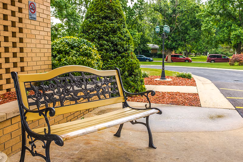 A decorative outdoor bench with a floral metal backrest is placed on a concrete patio next to a brick wall with a no smoking sign. Surrounding the area are green bushes, trees, a lamp post, and a parking lot with several parked cars in the background.