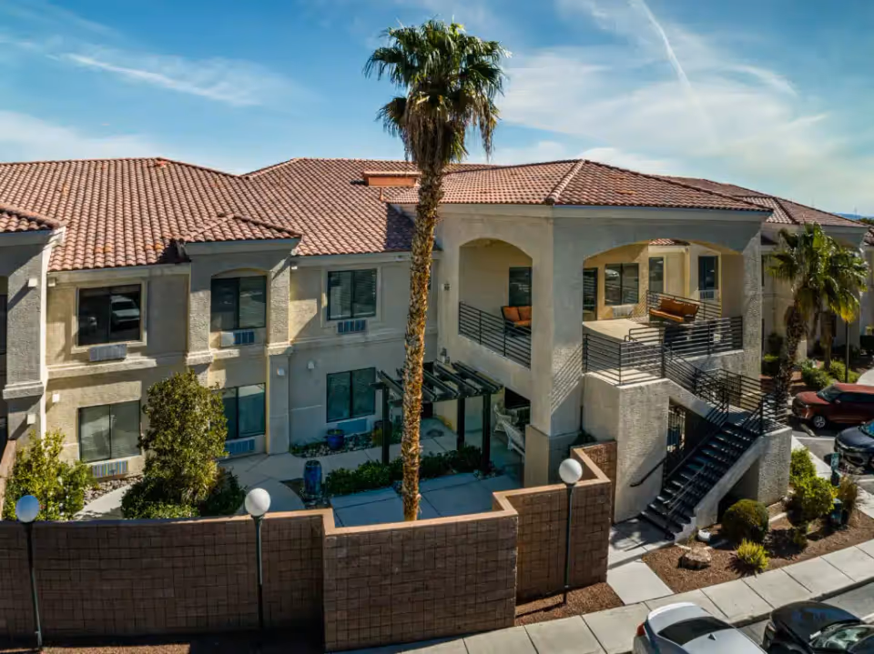 Exterior view of a two-story Mediterranean-style senior living building with a red tile roof, exterior stairway, balconies, palm trees, and parked cars.