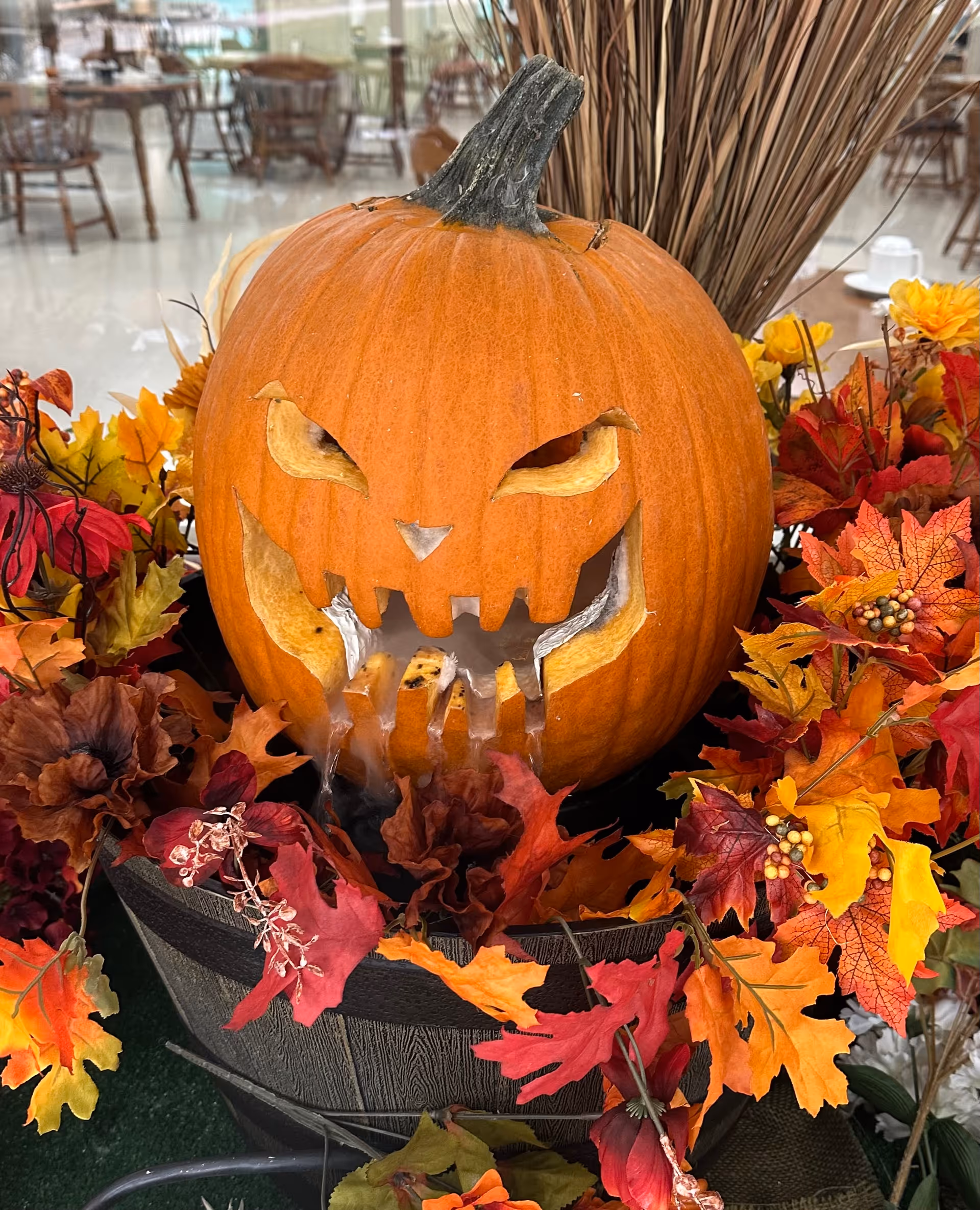 A carved pumpkin with a menacing face, featuring sharp teeth and slanted eyes, surrounded by colorful autumn leaves and flowers in a wooden barrel. The background shows a dining area with tables and chairs.