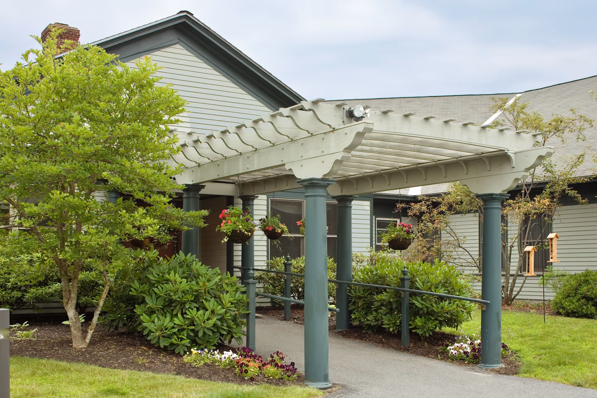 Outdoor view of a senior living facility entrance with a white pergola supported by green columns. There are hanging flower baskets and well-maintained shrubs and trees surrounding the pathway leading to the building.