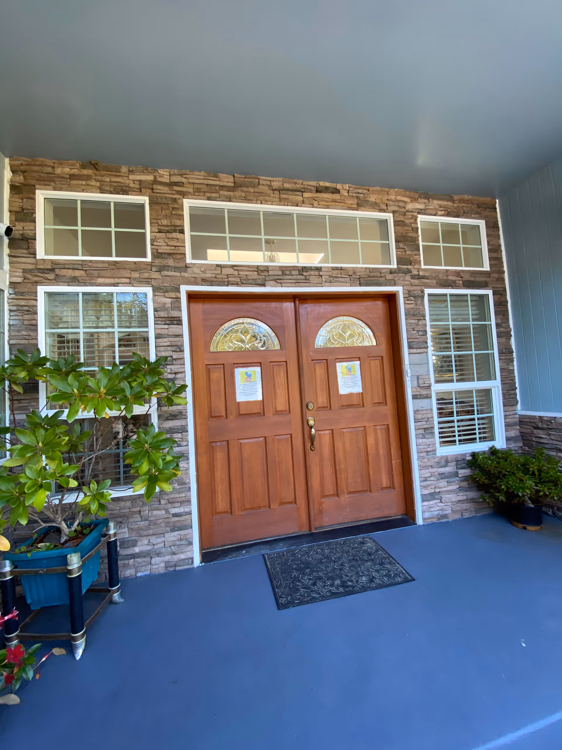 Front entrance of a building with double wooden doors featuring decorative glass panels. The doors are set in a stone facade with multiple rectangular windows above and on either side. There are potted plants on both sides of the entrance and a black doormat on the blue floor in front of the doors.