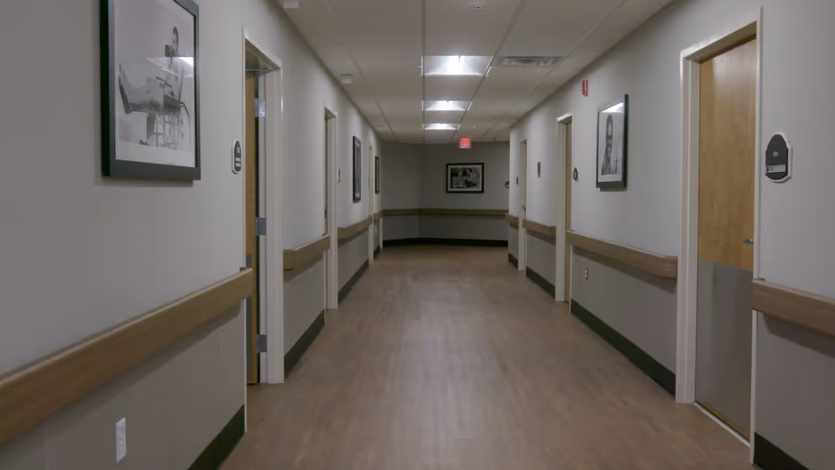 A long, empty hallway in a healthcare facility with wooden doors on both sides, beige walls with wooden handrails, framed black and white photographs on the walls, and overhead fluorescent lighting.