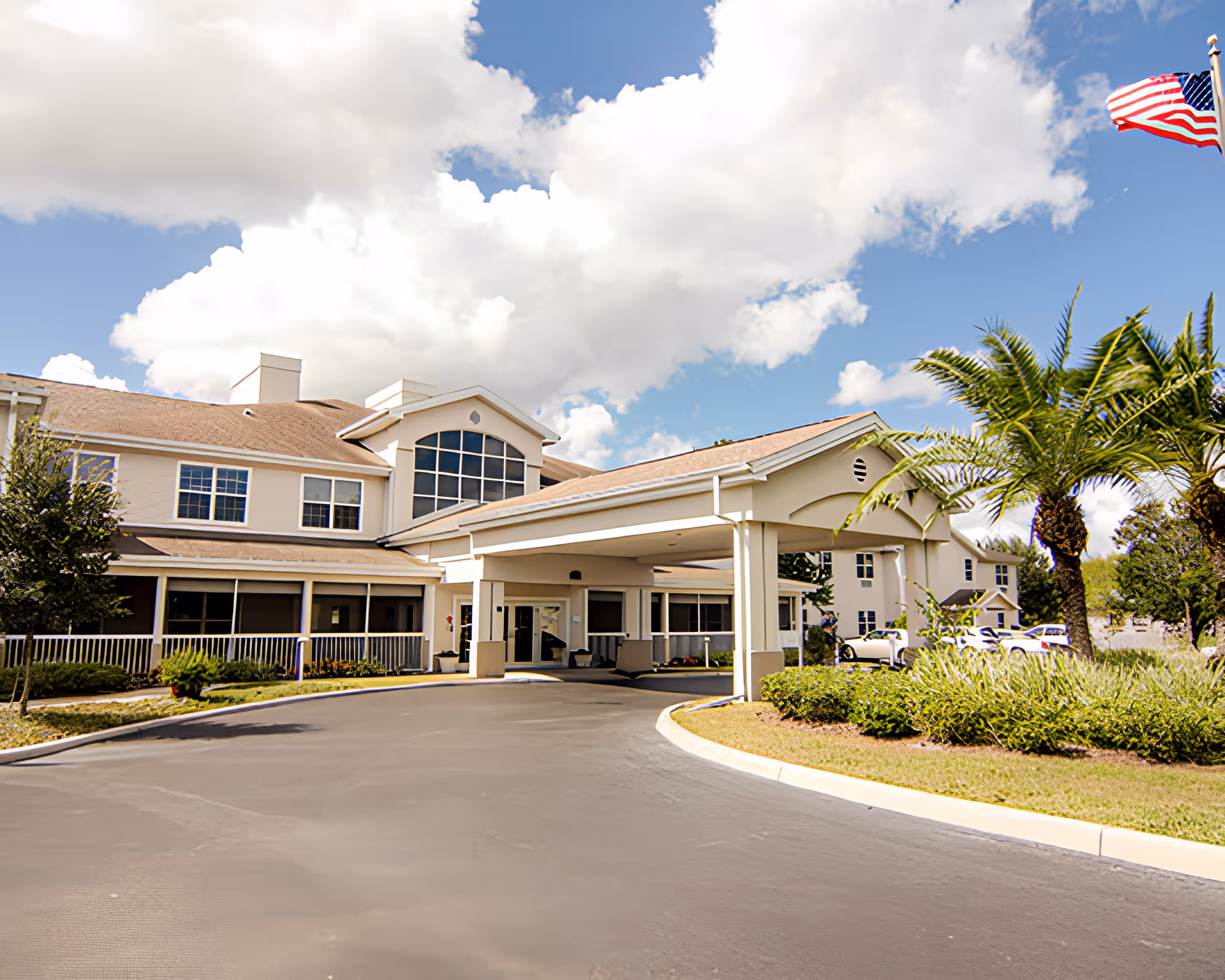 Exterior view of a senior living facility named Brookdale Highlands with a covered entrance, palm trees, and an American flag flying on a sunny day with partly cloudy skies.
