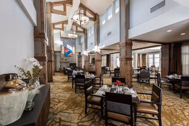 A spacious dining room with high vaulted ceilings featuring exposed wooden beams and large hanging light fixtures. The room is furnished with multiple dark wood tables and chairs, each set with white napkins, silverware, and small flower arrangements. The carpet has a patterned design, and there is a fireplace with a colorful abstract painting above it at the far end of the room. Large windows allow natural light to fill the space.
