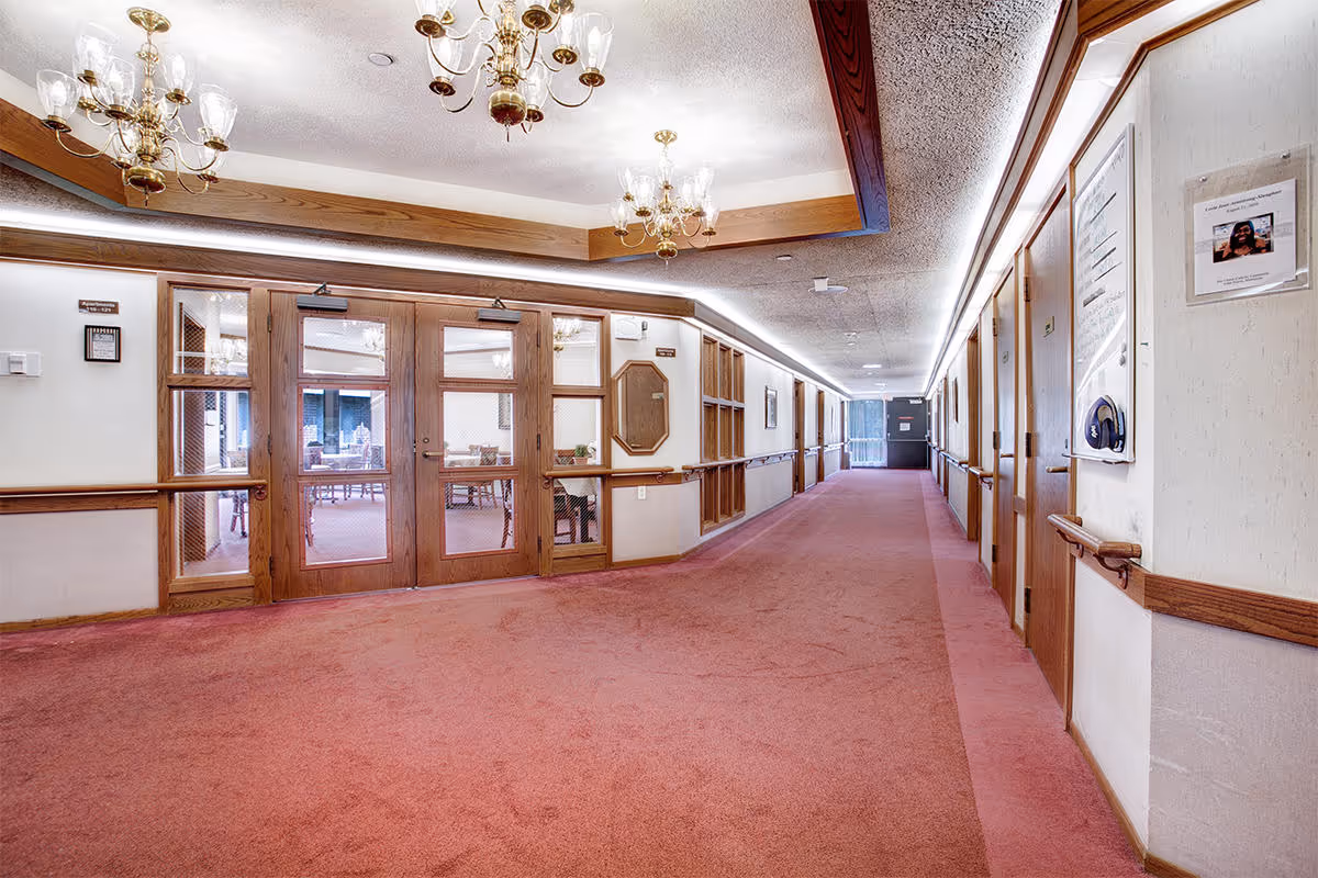 A long indoor hallway with red carpet and wooden handrails on both sides. The ceiling has recessed lighting and two brass chandeliers. On the left side, there are wooden framed glass doors leading to a room with tables and chairs. On the right side, there are several closed wooden doors and a bulletin board with notices. The hallway extends to a door at the far end.
