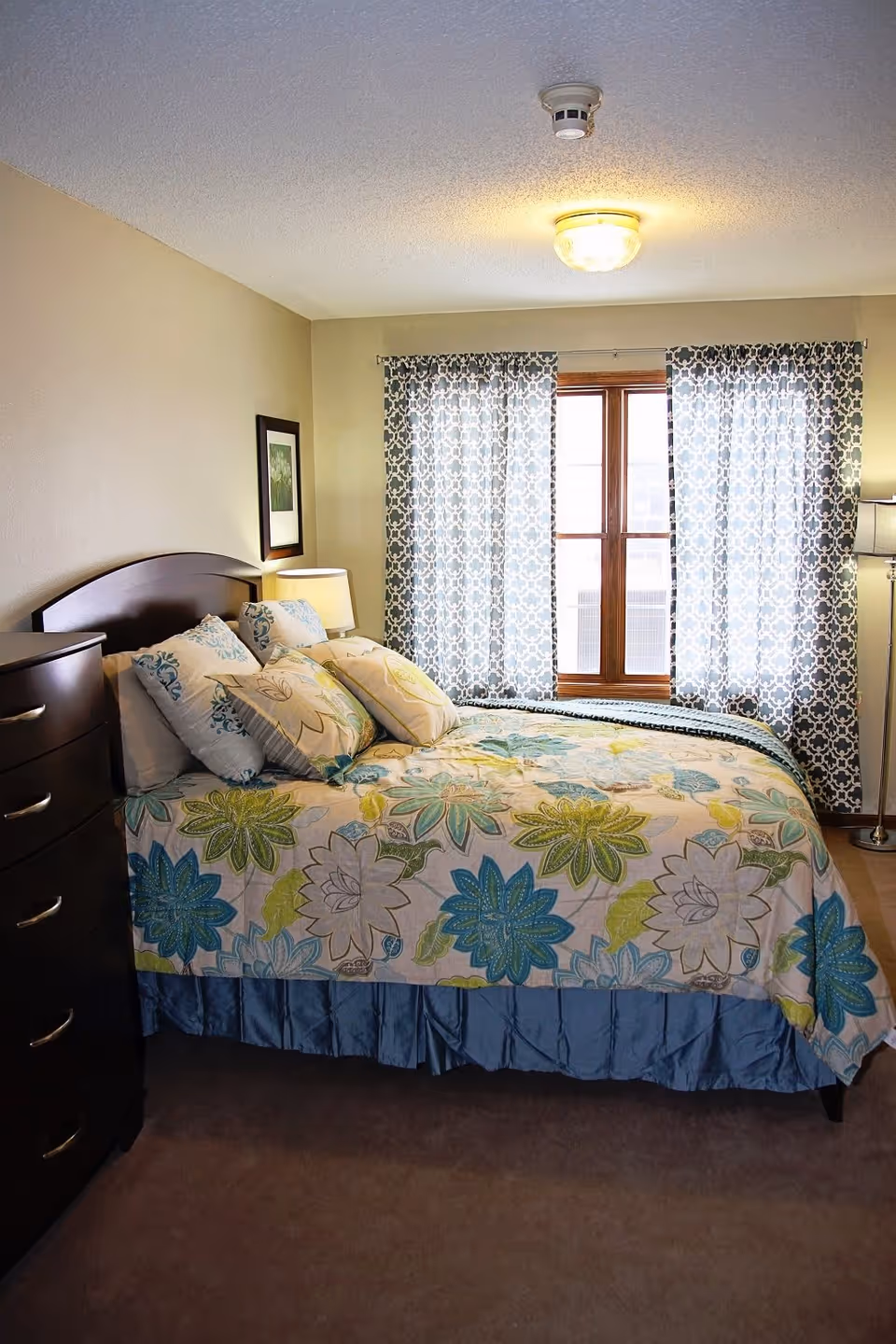A cozy bedroom with a bed covered in a floral patterned quilt in shades of blue, green, and yellow. The bed has multiple pillows and is positioned against a beige wall. There is a dark wooden dresser to the left and a window with patterned curtains letting in natural light. A framed picture hangs on the wall above a bedside lamp, and a floor lamp stands in the corner.