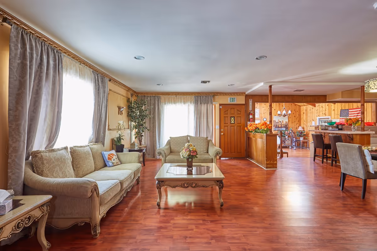 A spacious senior living facility common area with wooden flooring, beige upholstered sofas, a decorative coffee table with a flower arrangement, large windows with curtains, and a wooden door with an exit sign above it. The room extends into a dining area with chairs, tables, and an American flag on the wall.