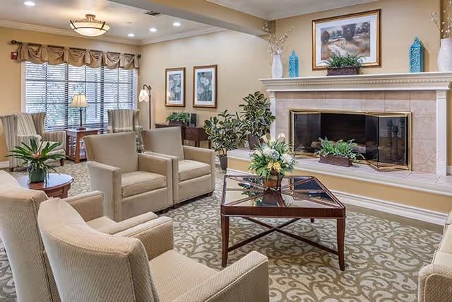 A cozy living room with beige armchairs arranged around a glass coffee table with a floral centerpiece. The room features a fireplace with decorative vases and a framed painting above it. There are large windows with curtains, framed artwork on the walls, and several potted plants adding greenery to the space.