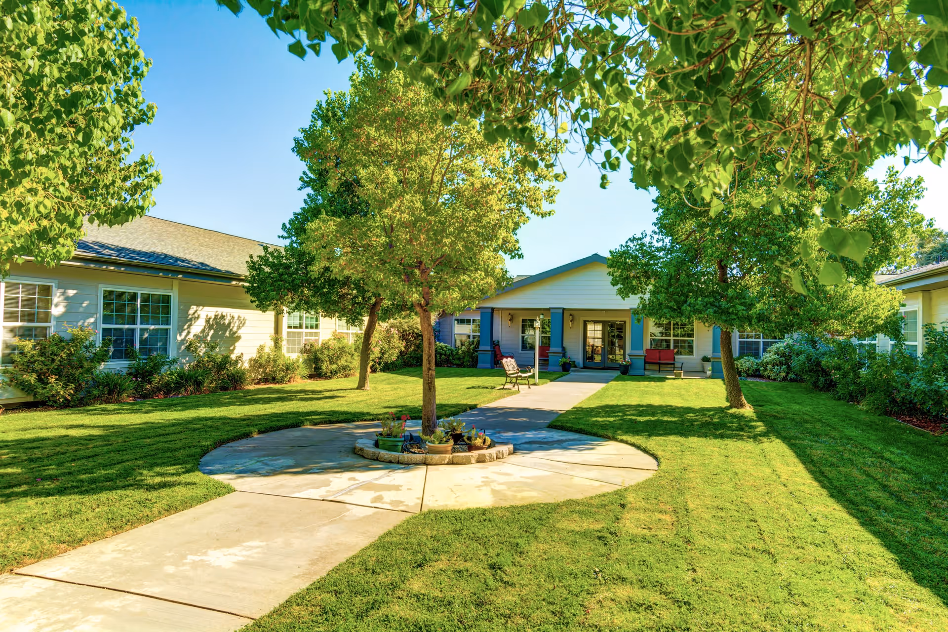 A sunny outdoor courtyard area at a senior living facility with a paved walkway leading to a building entrance. The courtyard features green grass, several trees, a circular flower bed with potted plants, and benches near the entrance. The building has light-colored siding and multiple windows.