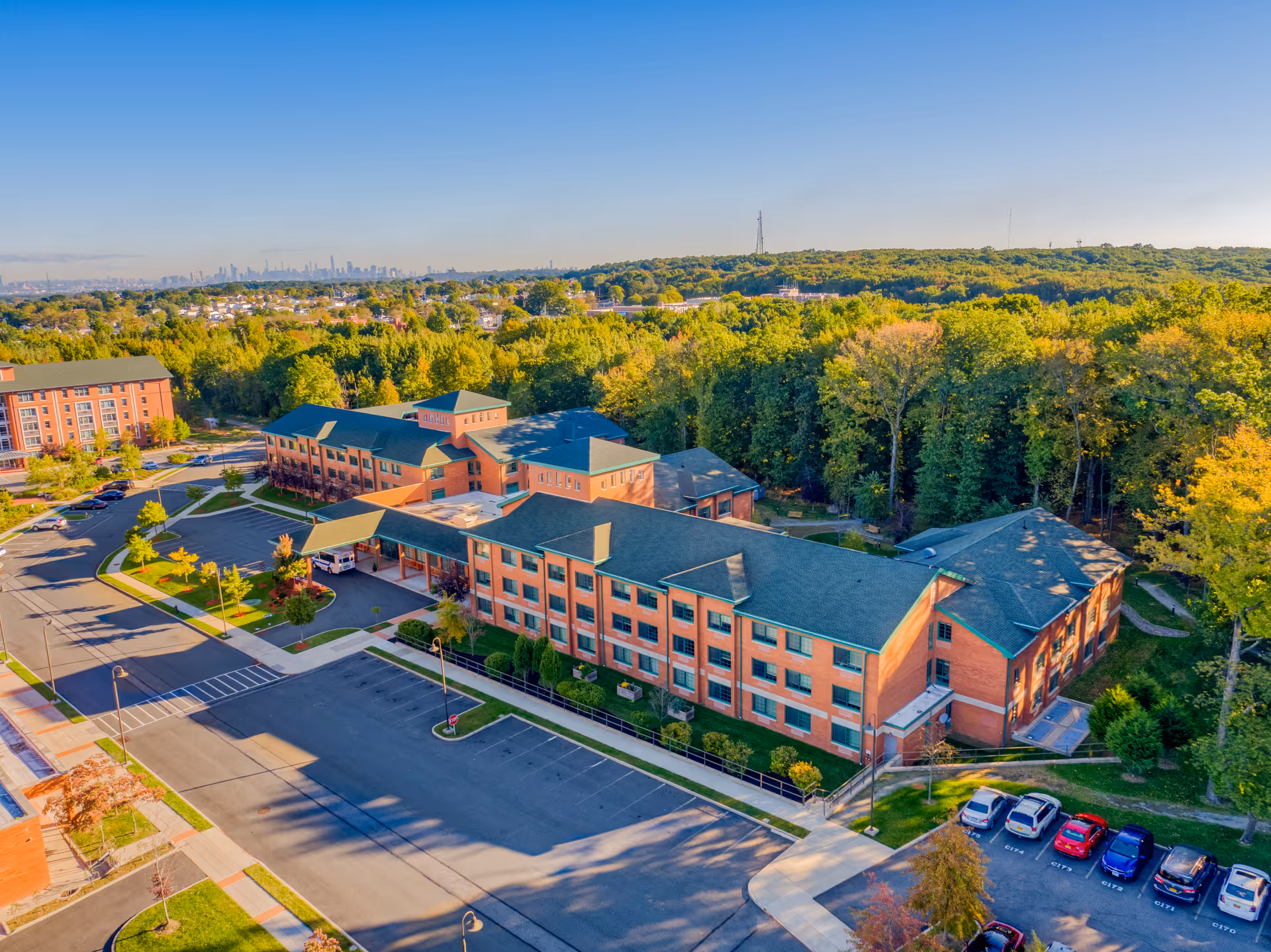 Aerial view of The Brielle senior living facility surrounded by trees and greenery, with a parking lot and several cars visible. The building is a large, multi-story brick structure with green roofs, situated near a wooded area under a clear blue sky.