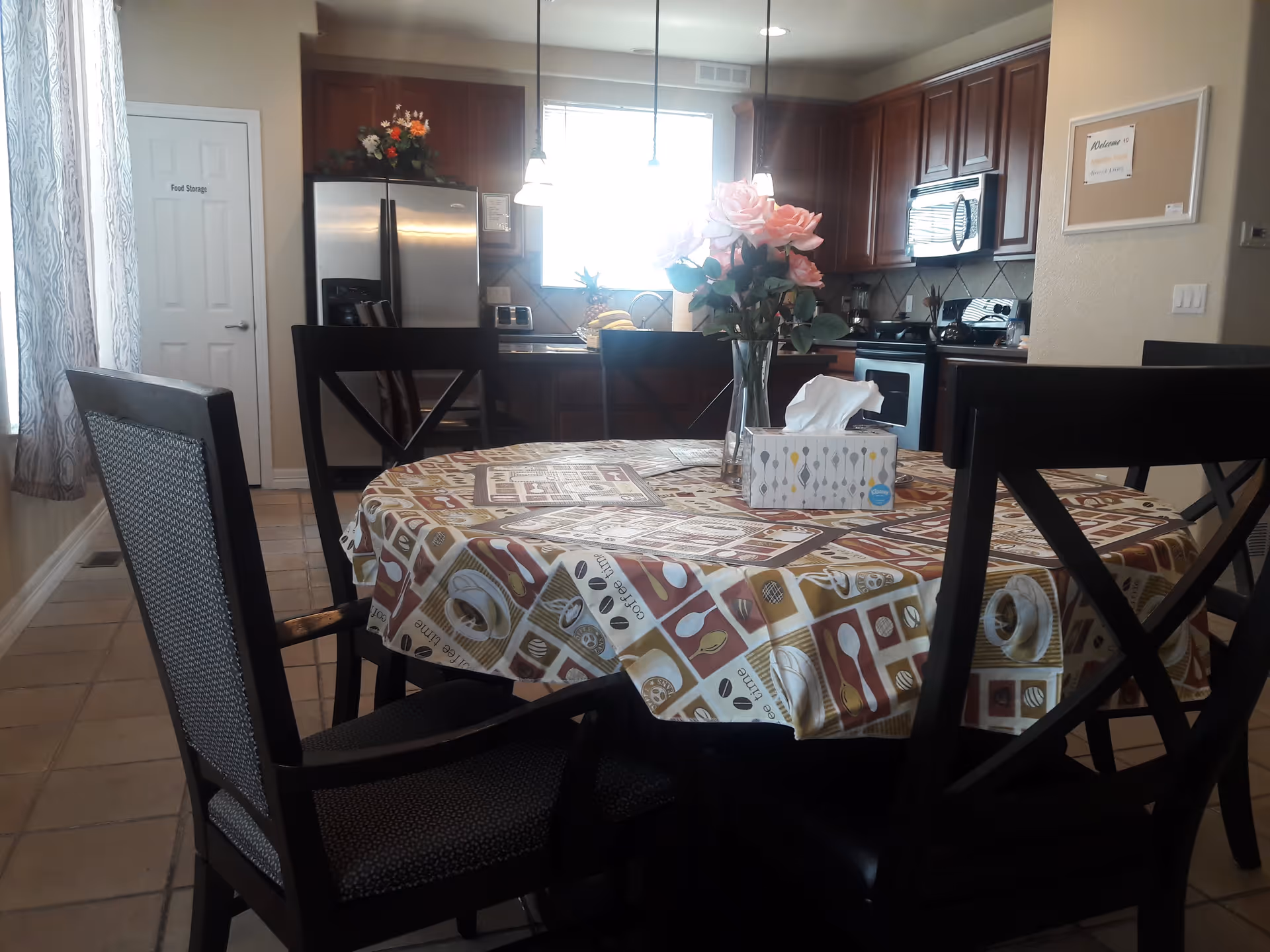 A dining area with a round table covered by a patterned tablecloth featuring coffee and kitchen-themed designs. On the table, there is a vase with pink flowers and a box of tissues. Surrounding the table are four dark wooden chairs with cushioned seats. In the background, there is a kitchen with dark wooden cabinets, a stainless steel refrigerator, a microwave, and a stove. A door labeled 'Food Storage' is visible to the left, and a window with patterned curtains lets in natural light.