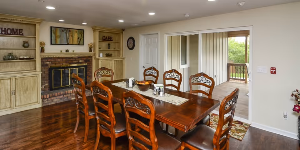 Dining room with a large wooden table surrounded by eight chairs with decorative backs. The room features hardwood flooring, built-in shelves labeled 'HOME' and 'CAFE', a brick fireplace with a glass door, and a sliding glass door leading to an outdoor porch area with greenery visible outside.