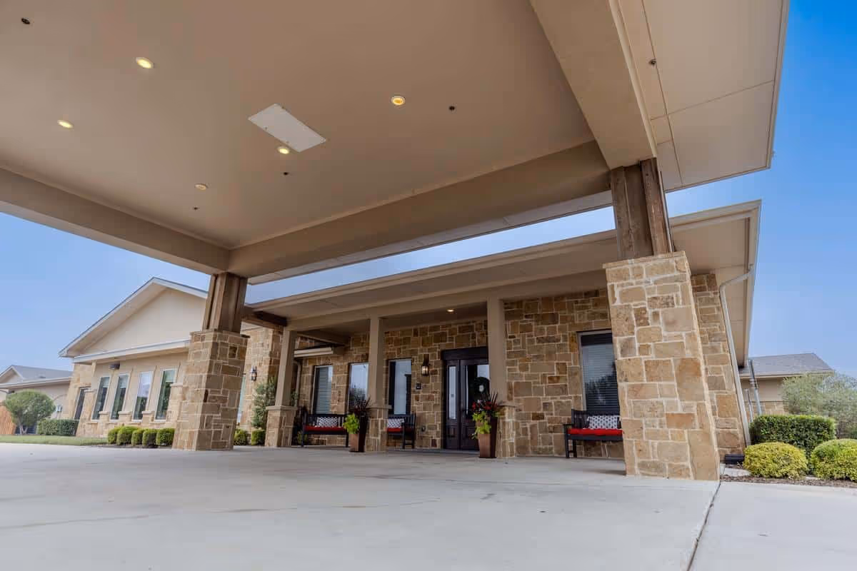 Entrance area of a building with stone pillars and walls, a covered driveway, benches with cushions, potted plants, and a clear blue sky.