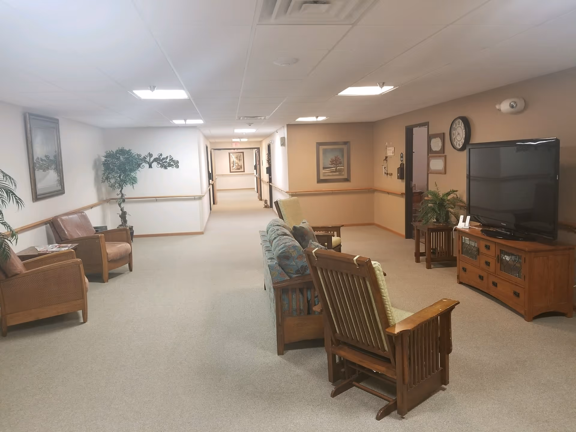 A quiet hallway in a retirement community with beige walls and carpet. There are several wooden chairs with cushions arranged facing a large flat-screen TV on a wooden cabinet. The hallway is decorated with framed pictures, a clock, and potted plants. The ceiling has recessed lighting and ventilation panels.