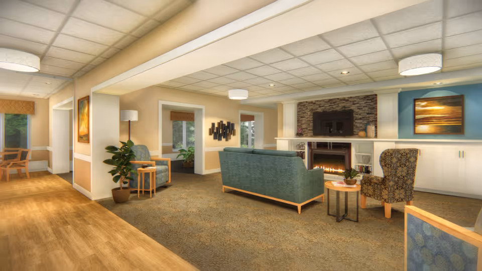 A cozy senior living facility common area with a green sofa, patterned armchair, small round table with a plant, and a fireplace with a TV mounted above it. The room has beige walls, carpeted flooring, and large windows with brown valances letting in natural light. There are also decorative wall art pieces and potted plants.