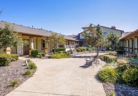 Sunny landscaped courtyard with a paved walkway, chairs, and surrounding assisted-living buildings.