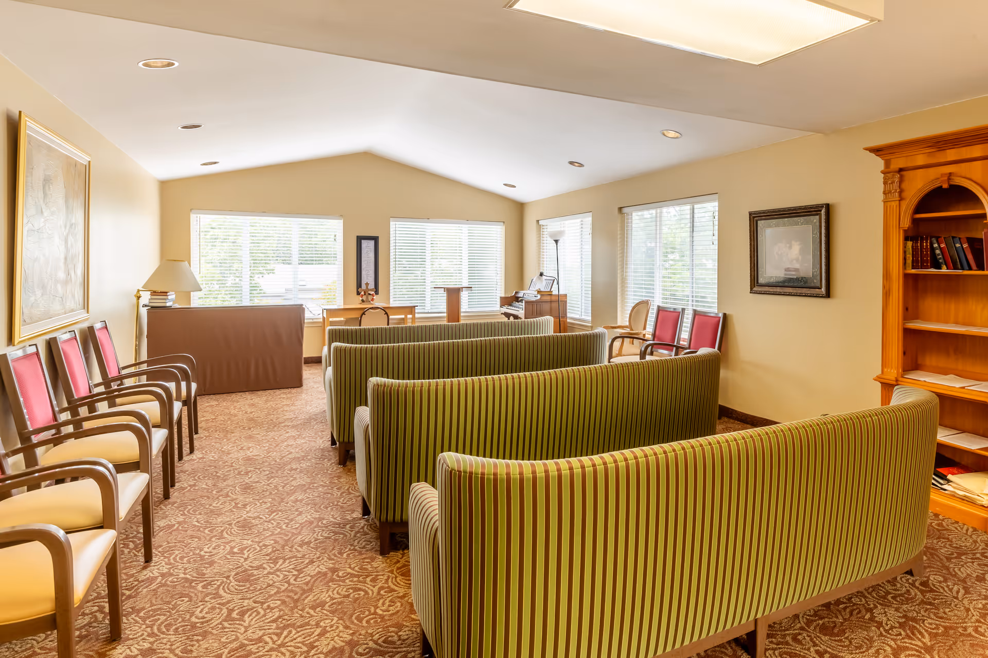 A bright room with large windows covered by white blinds, featuring three green and brown striped sofas arranged in rows facing a small wooden podium and an organ. There are several chairs with red cushions along the walls, a wooden bookshelf with books, a floor lamp, and framed artwork on the walls. The room has a patterned carpet and recessed lighting in the ceiling.