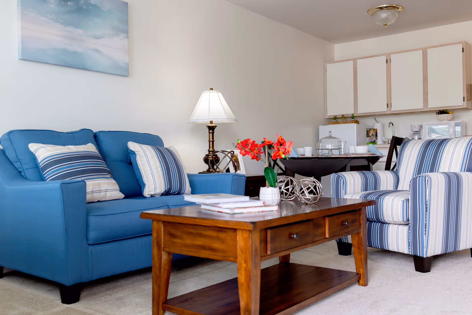 A cozy living room area featuring a blue sofa with striped pillows, a matching striped armchair, a wooden coffee table with books and a small vase of red flowers, a table lamp, and a small kitchenette with white cabinets and appliances in the background.