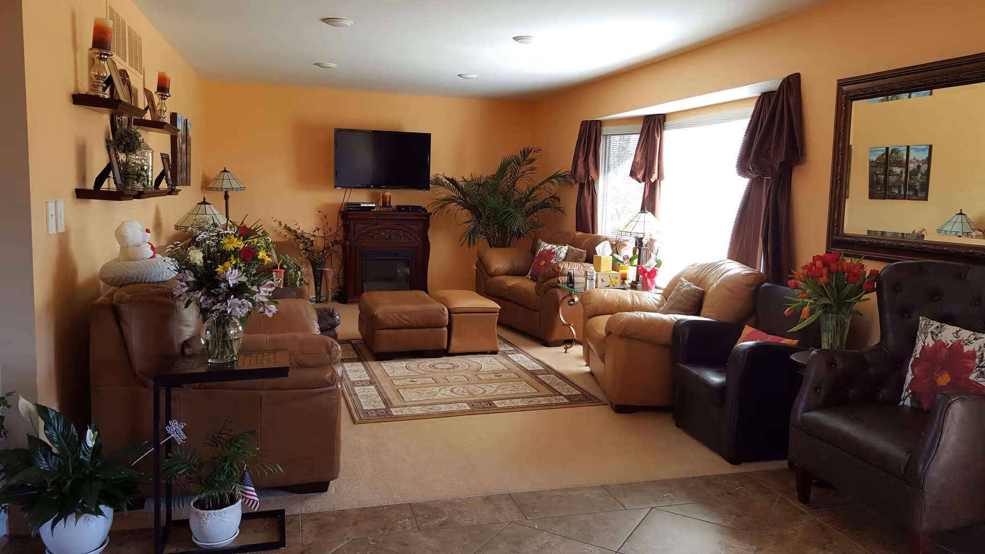 A cozy living room with beige walls and carpet, featuring multiple leather armchairs and sofas arranged around a patterned area rug. A flat-screen TV is mounted on the wall above a wooden electric fireplace. The room is decorated with various plants, flowers in vases, a large mirror on one wall, and curtains framing a large window letting in natural light.