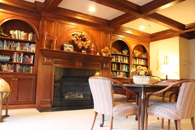 Cozy common room with wood-paneled built-in bookshelves and a fireplace, plus a round table and upholstered chairs.