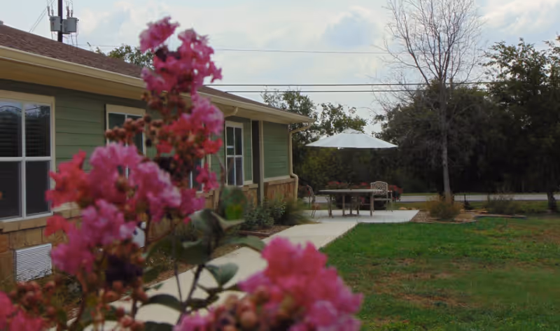 Pink flowering shrub in the foreground with a green cottage, walkway and an outdoor patio table with umbrella on a lawn.