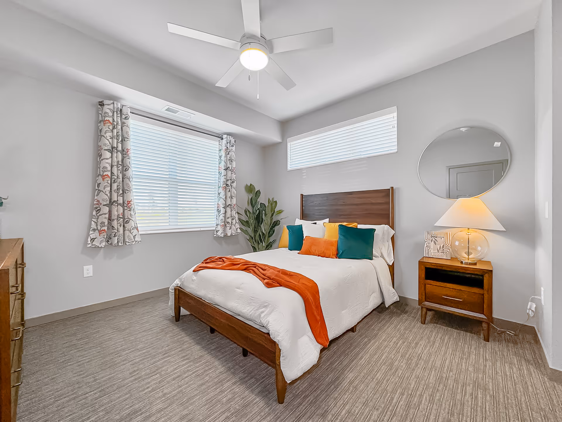 A bright bedroom with a wooden bed frame and white bedding accented by colorful pillows and an orange throw. The room features two windows with blinds, one with floral curtains, a ceiling fan with a light, a wooden nightstand with a lamp and decorative items, a round mirror on the wall, and a green plant in the corner.