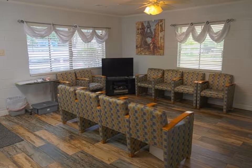 A senior living facility common area with patterned armchairs arranged in two rows facing a flat-screen TV on a black stand. The room has wood-style flooring, white walls, two windows with white valance curtains, a ceiling fan with a light, and a painting on the wall.