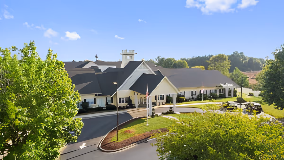 Front exterior of a single-story assisted living building with a covered porte-cochère, circular driveway, and surrounding trees.