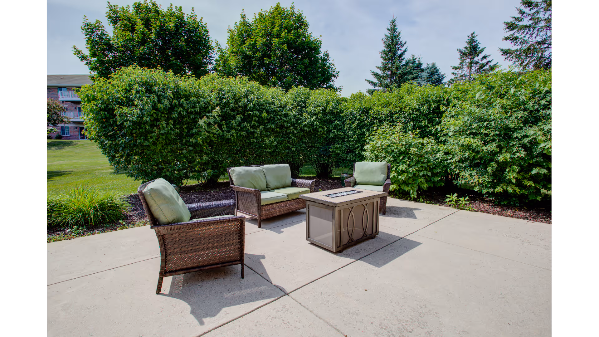 Outdoor patio area with three wicker chairs and a loveseat, all with green cushions, arranged around a rectangular fire pit table. The patio is surrounded by lush green bushes and trees under a partly cloudy sky.