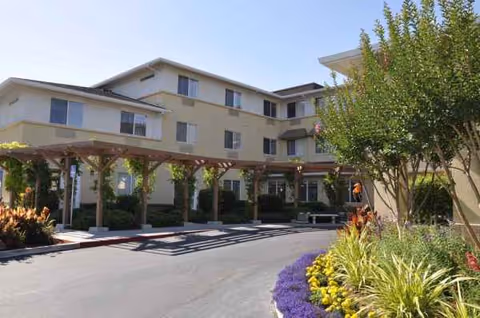 Exterior view of Carlton Senior Living San Jose: beige building, pergola walkway, and landscaped garden.