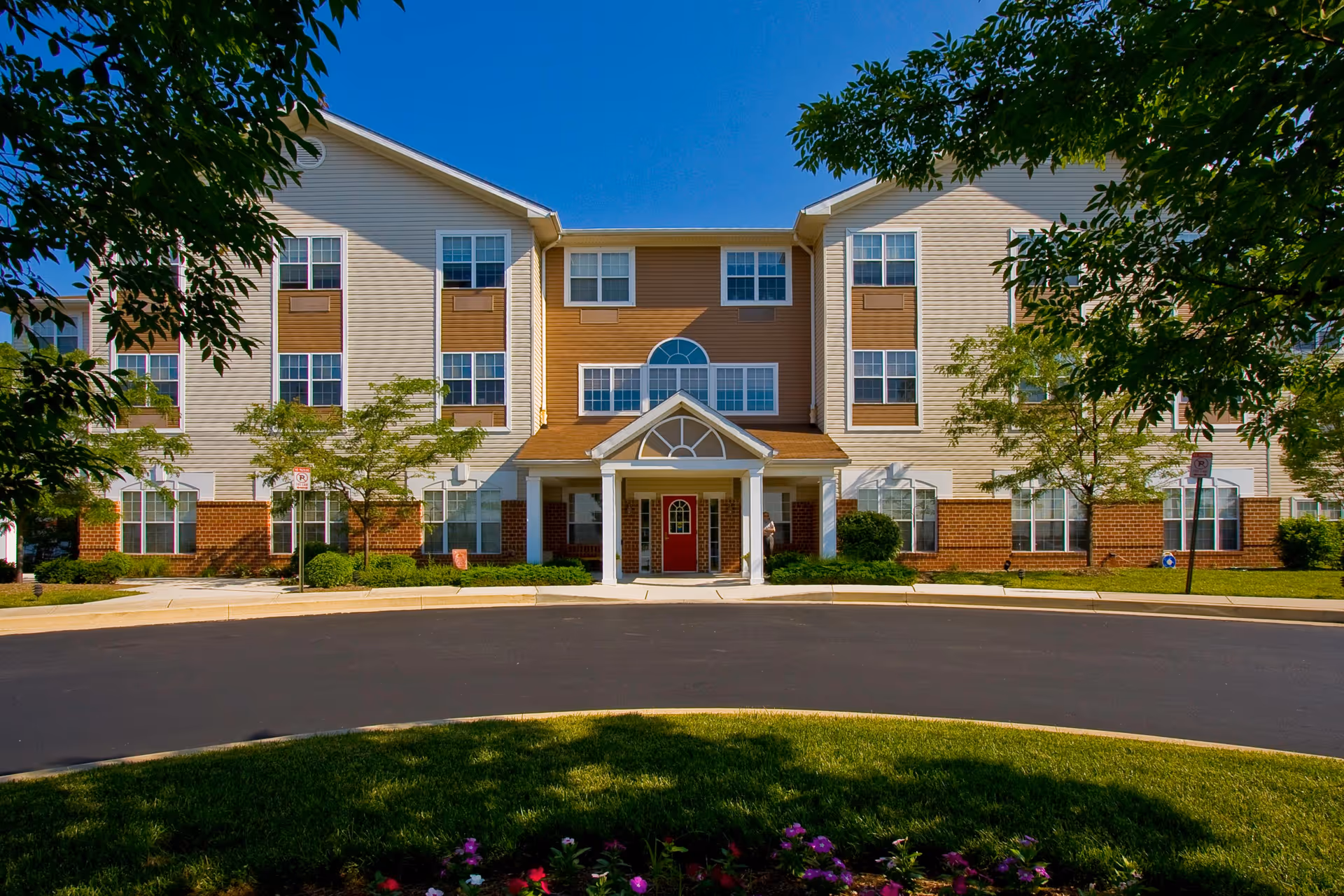 Front exterior of a three-story senior living building with a central covered entrance, red door, landscaped grounds and a circular driveway.