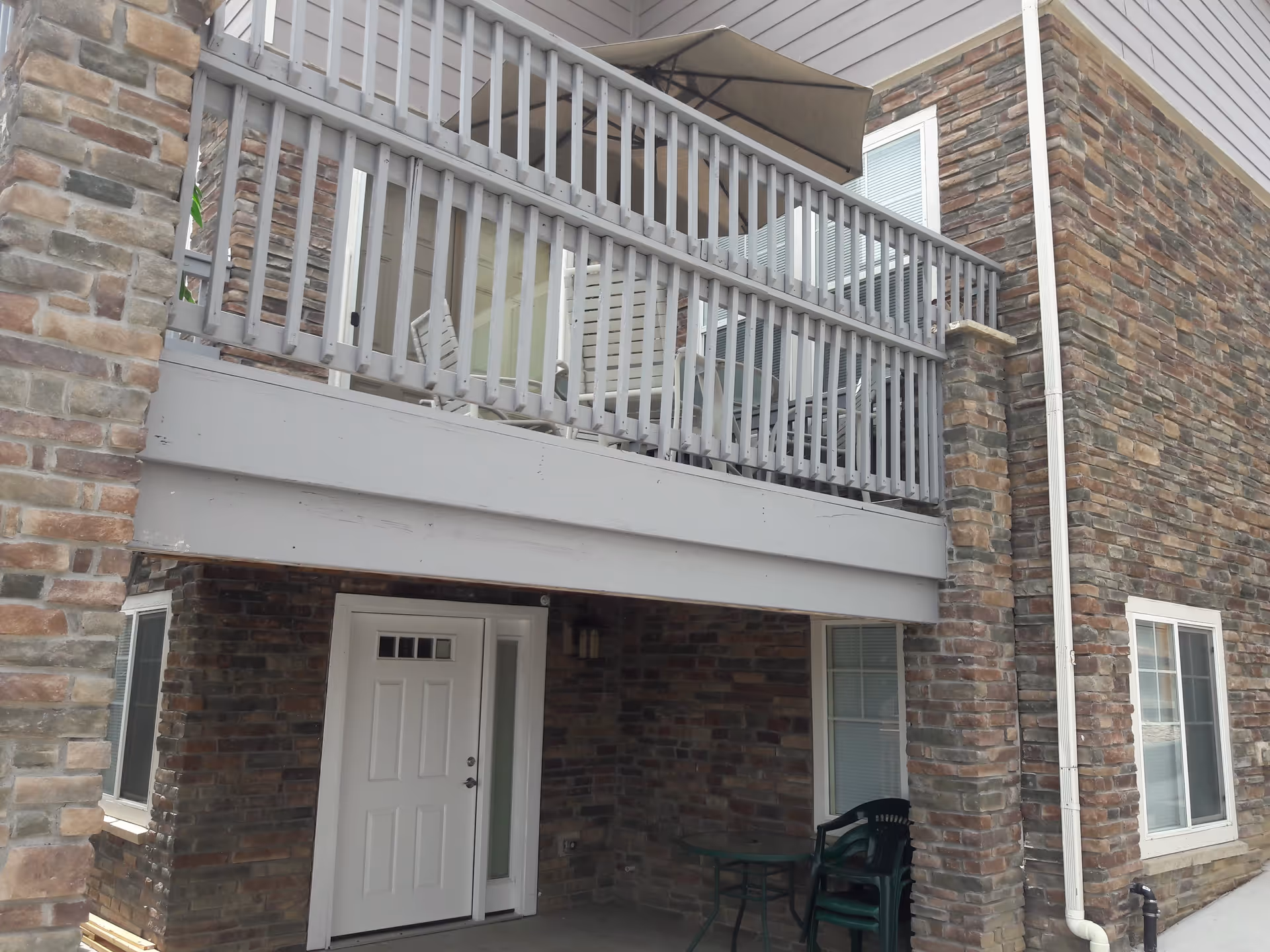 Exterior view of a building corner with stone and siding walls, featuring a white door under a covered patio area and a balcony above with a railing, patio furniture, and an umbrella.