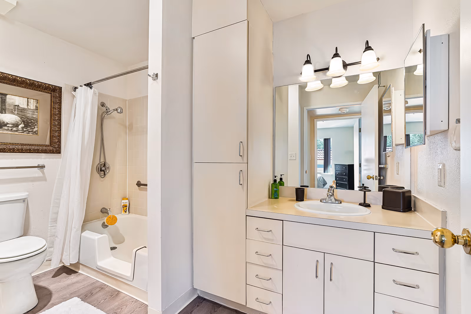 A clean and well-lit bathroom featuring a white toilet, a bathtub with a built-in seat and a showerhead with a white shower curtain. There is a large mirror above a white vanity with multiple drawers and cabinets, a sink, and countertop accessories including soap dispensers and a tissue box. A framed picture hangs on the wall above the toilet, and the bathroom floor has wood-like flooring with a white rug.