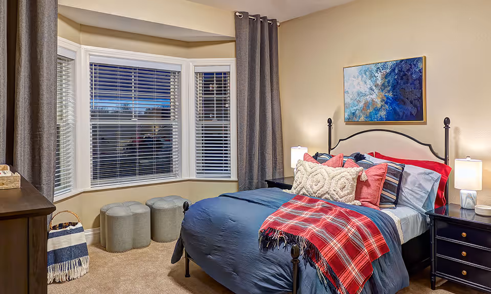 A cozy bedroom with a bed dressed in blue bedding, multiple decorative pillows, and a red plaid throw blanket. The room features a large bay window with white blinds and gray curtains, two small gray ottomans, a wooden dresser, two black nightstands with lamps, and a blue and white abstract painting on the beige wall.